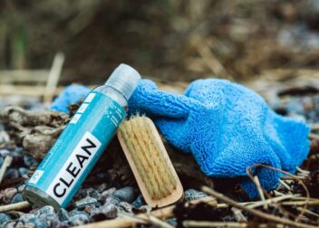 Bottle labeled “CLEAN,” wooden brush, and blue microfiber cloth on pebbles outdoors.