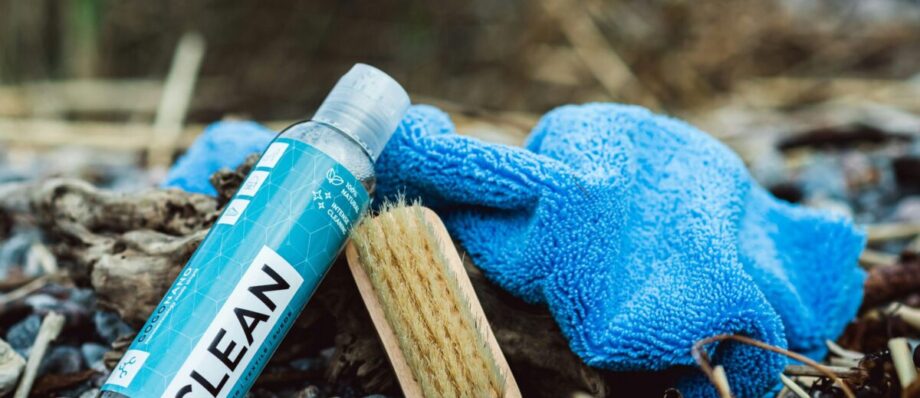 Bottle labeled “CLEAN,” wooden brush, and blue microfiber cloth on pebbles outdoors.