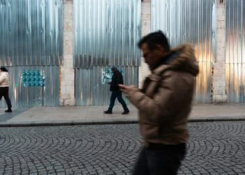 Three people walk on a cobblestone street by a corrugated metal wall; one in the foreground is blurred and using a phone.