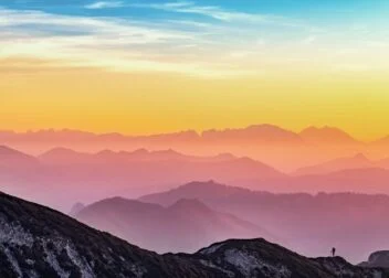 Person on a mountain ridge at sunrise, viewing layers of misty mountains beneath a colorful sky.