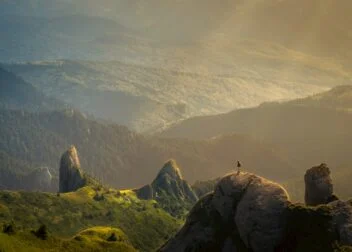 Person standing on a rocky peak, overlooking forests and mountains in golden sunlight.