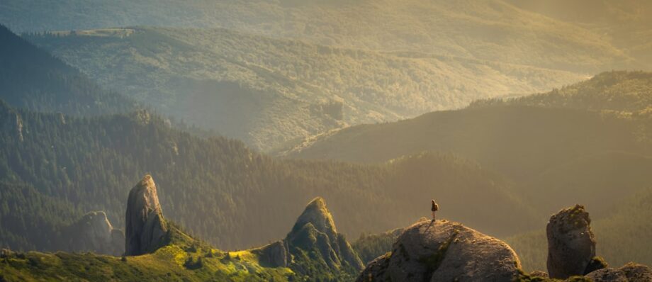 Person standing on a rocky peak, overlooking forests and mountains in golden sunlight.